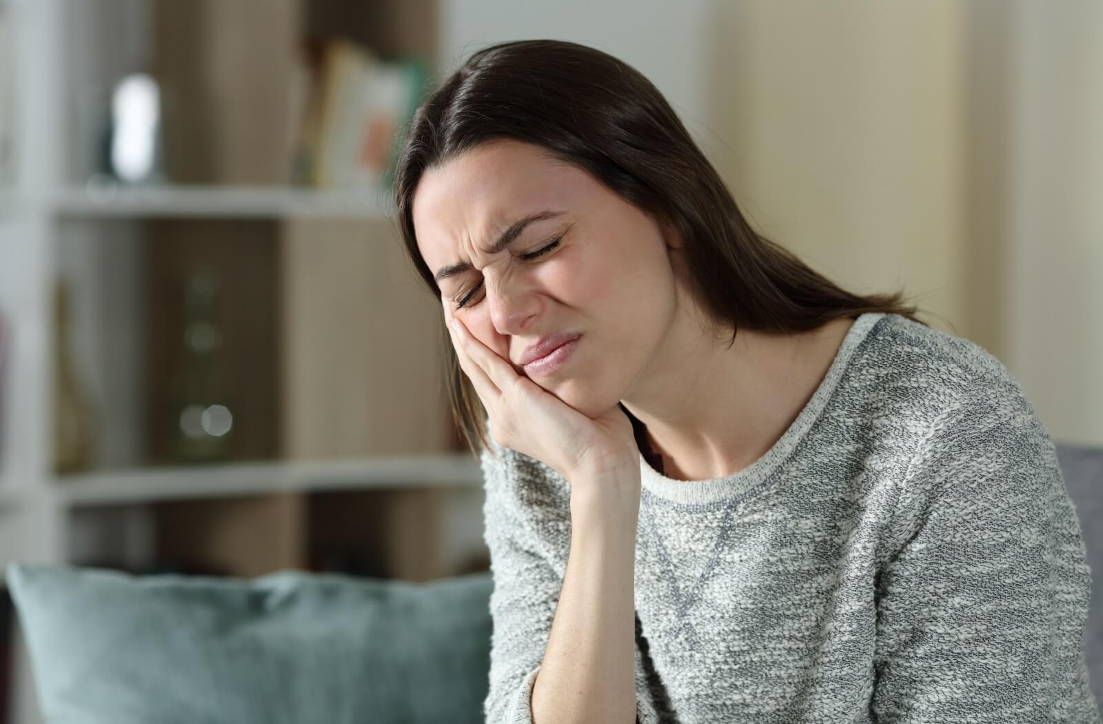 A woman sitting on her couch at home rubbing her jaw due to discomfort from her TMJ disorder.