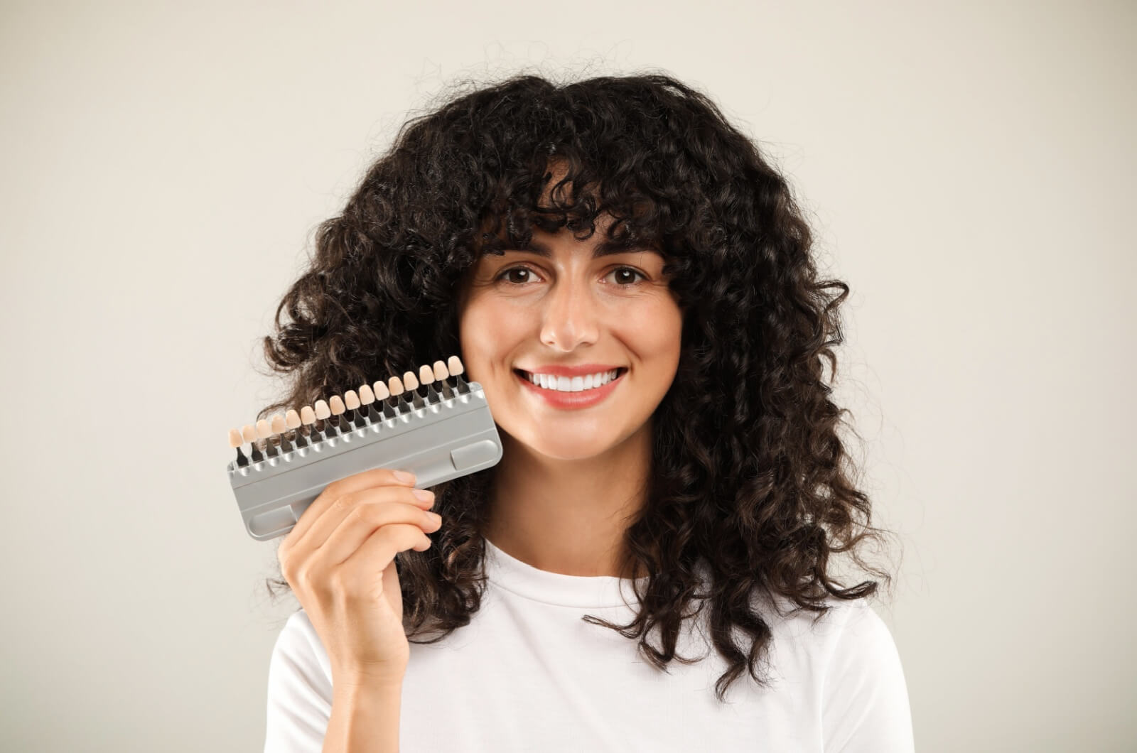 A smiling woman holding up a sample of dental veneers in various colours.