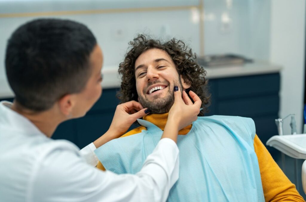 A smiling patient sitting in a dental chair while a dentist checks his teeth.