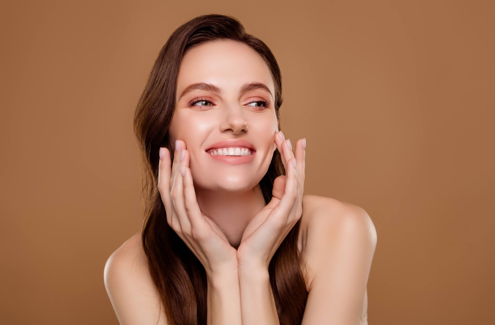 Smiling young woman with clear skin touching her face, posing against a neutral brown background.