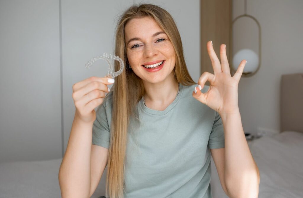 A smiling person holding up the clear Invisalign aligners and giving an "OK" hand gesture, highlighting successful overbite correction treatment.